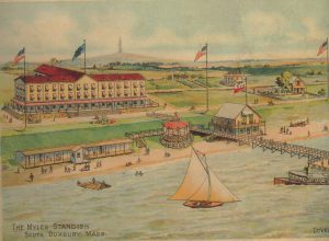 Large waterfront hotel with bathing houses, boats and dock, monument in background.