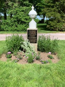 Pillar with plaque and finial in garden bed. 